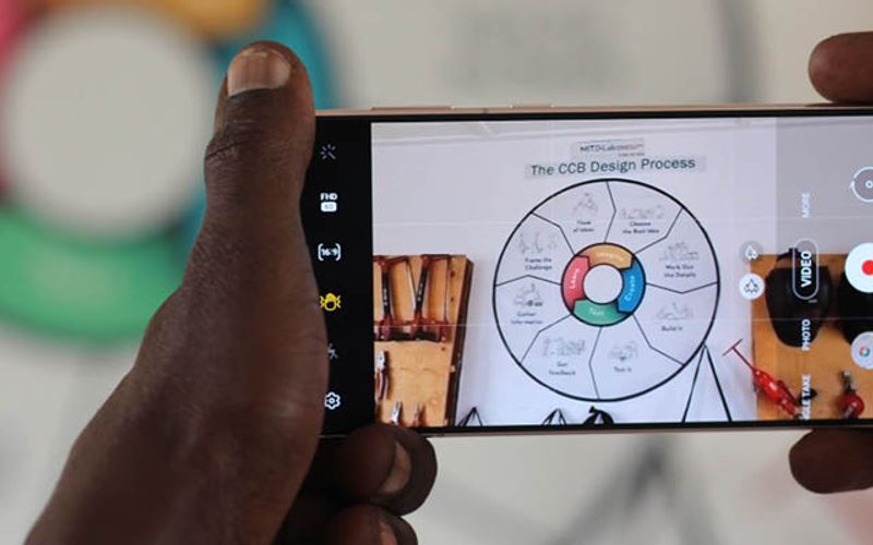 A person holds a smartphone and takes a photo of a diagram titled "The CCB Design Process," which shows a circular chart with labeled sections.